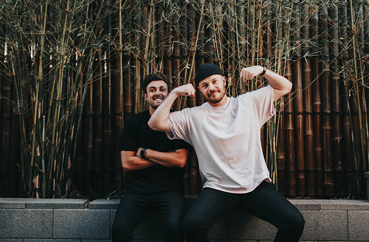 Two friends wearing beanies and smiling in front of a camera. Bamboo can be seen in the background.